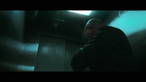 a bald dangerous looking man prepares a gun with the silencer in the elevator