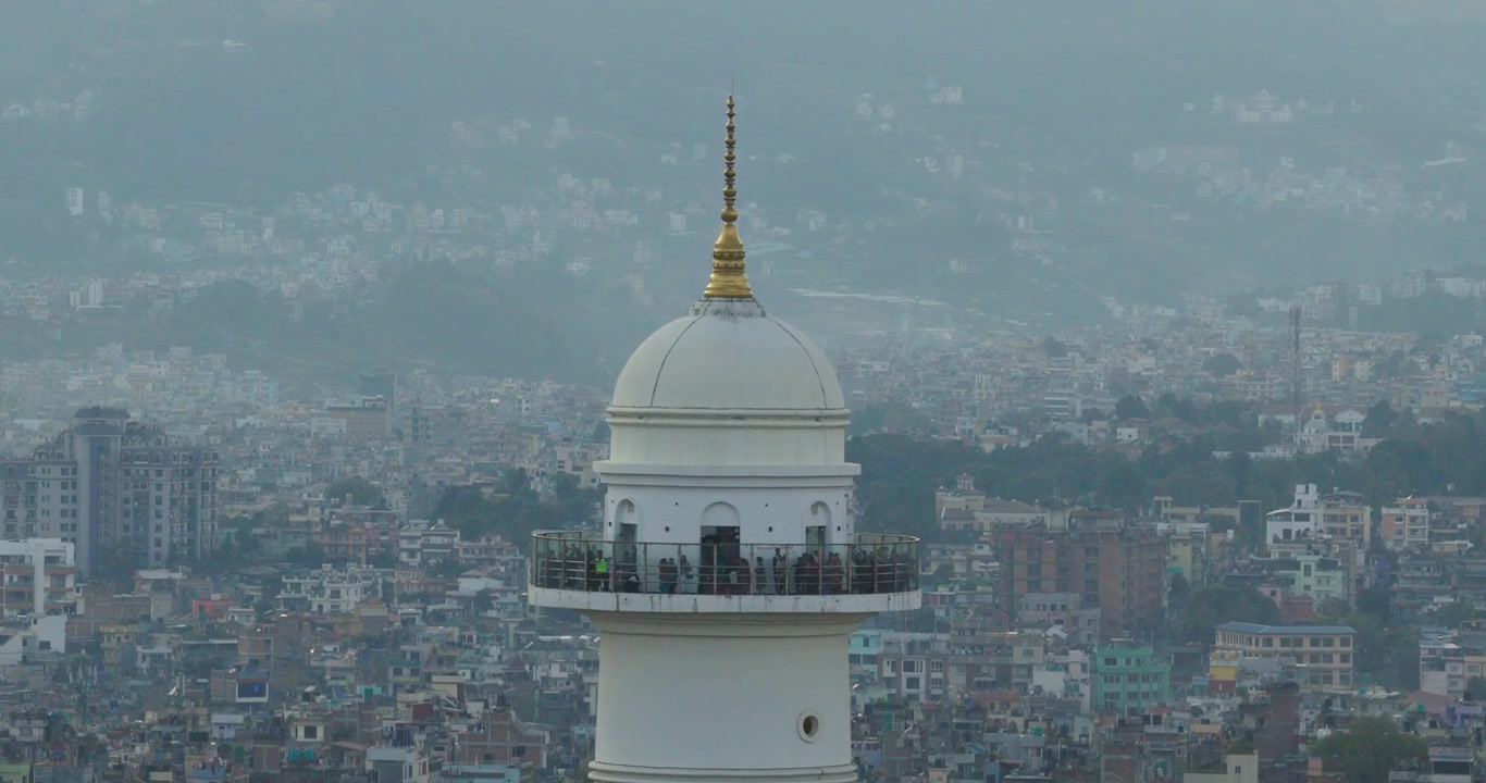 Dronen lähikuva Dharahara-tornista, joka näyttää sen huipun ja näköalatasanteen ihmisineen, taustalla vilkas Kathmandun kaupunki, Nepal ja kukkulat kultaisen tunnin aikana