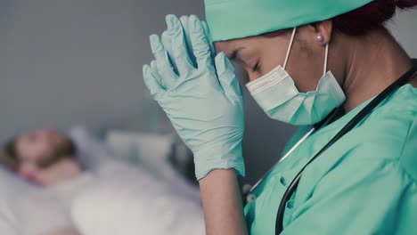 a young american female doctor praying next to a hospital bed