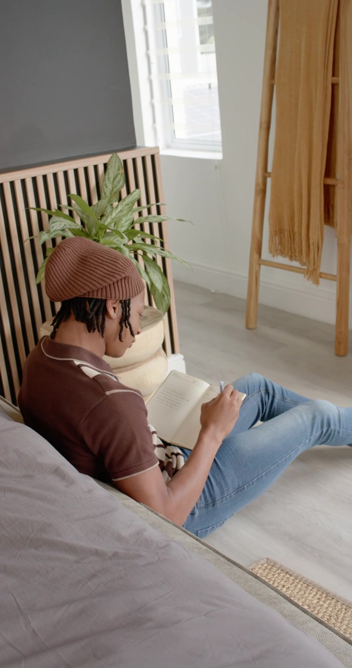 Video verticale: Uomo afroamericano seduto sul pavimento appoggiato al letto che legge un libro aperto per studiare