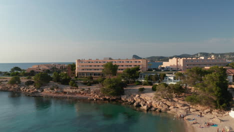 Luftaufnahme-Der-Menschenmenge,-Die-Sich-Am-Strand-Entspannen-Und-Genießen-Und-Im-Ozean-Schwimmen,-Umgeben-Von-Gebäuden-Und-Bergen-Auf-Ibiza-In-Spanien