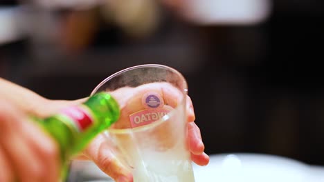 a hand pours beer from a green bottle into a glass, creating foam in a lively bar setting
