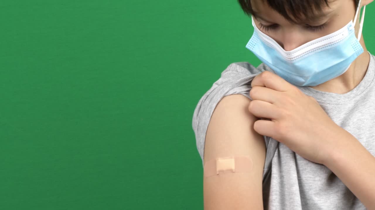 Teenage boy with medical protective mask showing to his arm with plaster after vaccination over green screen background. Vaccine and health care during pandemic Covid-19