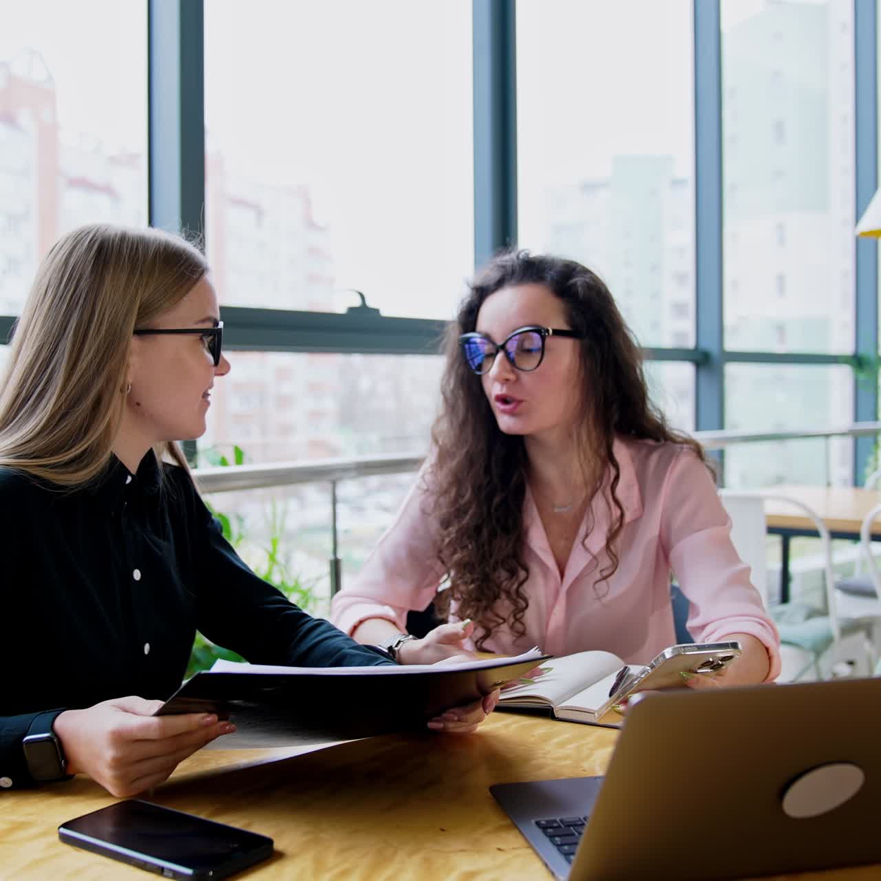 Young attractive women having conversation at office. Two ladies sit at desk having phone, documents and laptop in front of them