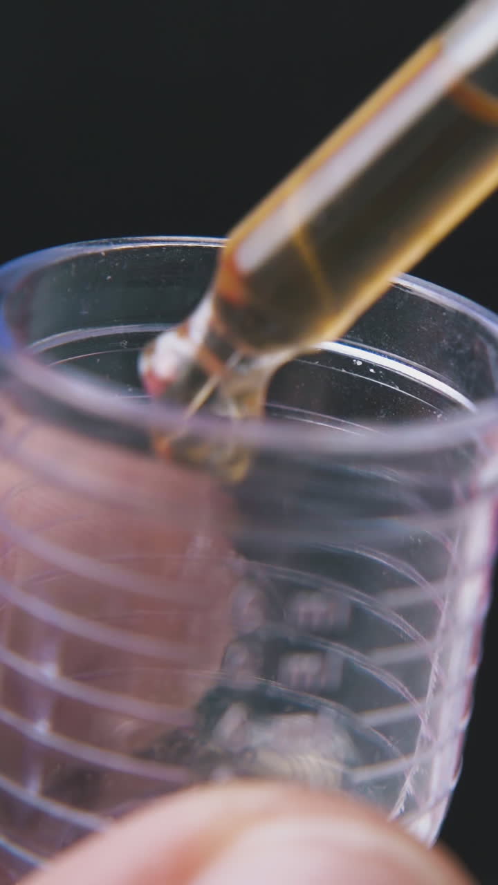lab assistant drops light brown water into small measuring cup with pipette on black background extreme close view