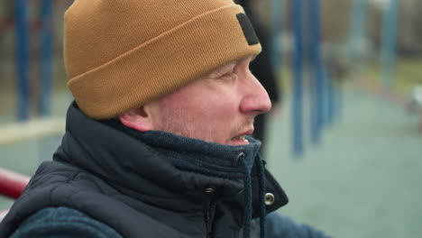 close-up of a coach sitting on a workout equipment, looking tired as he gazes at the boys playing football in the blurred background