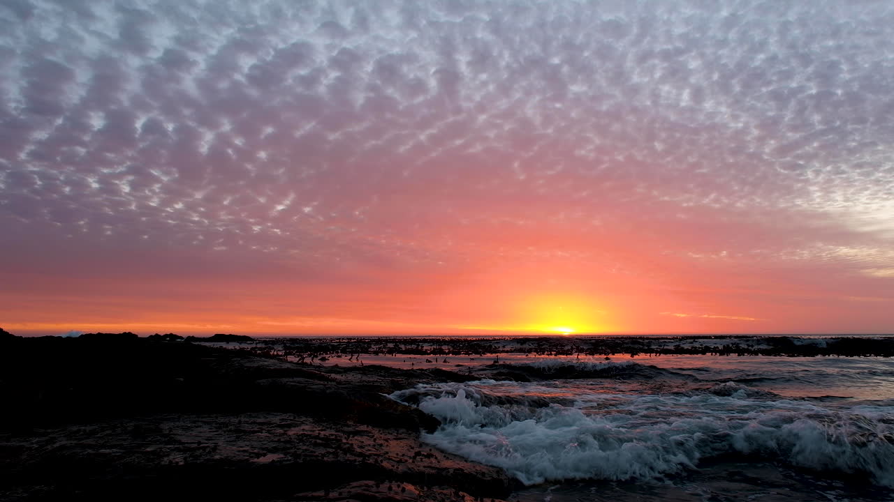 las suaves olas chocan contra las rocas, las nubes brillan de color naranja al atardecer sobre el océano
