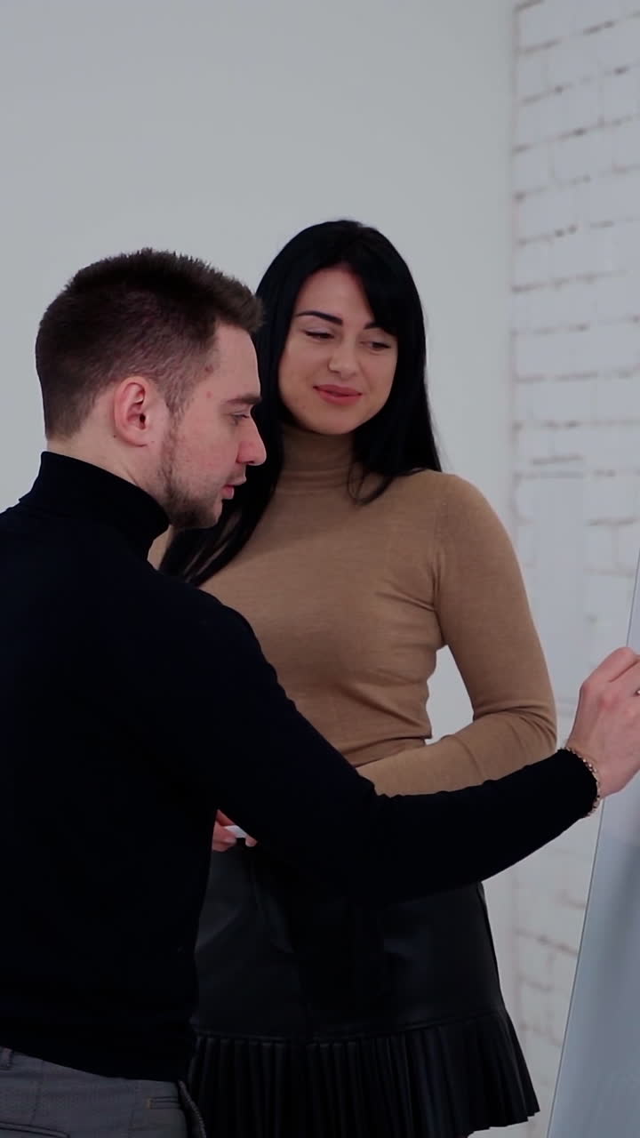 Young partners making new business plan. Handsome businessman is writing on a white board. Beautiful woman talking to a man in the office. Vertical video