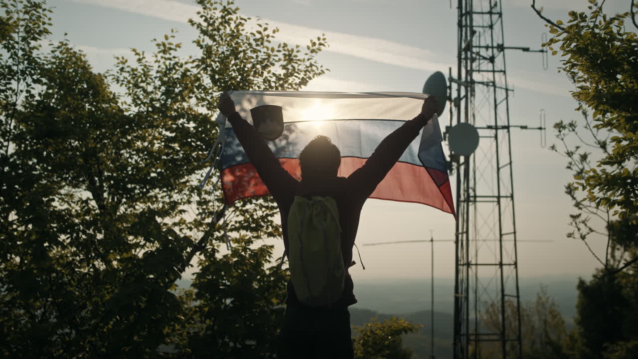 hombre que sostiene la bandera eslovena hacia el sol de verano cerca de los árboles y una torre de radio en la cima de la montaña slivnica