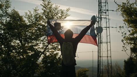 man holding slovenian flag towards the summer sun near trees and a radio tower on peak of mountain slivnica