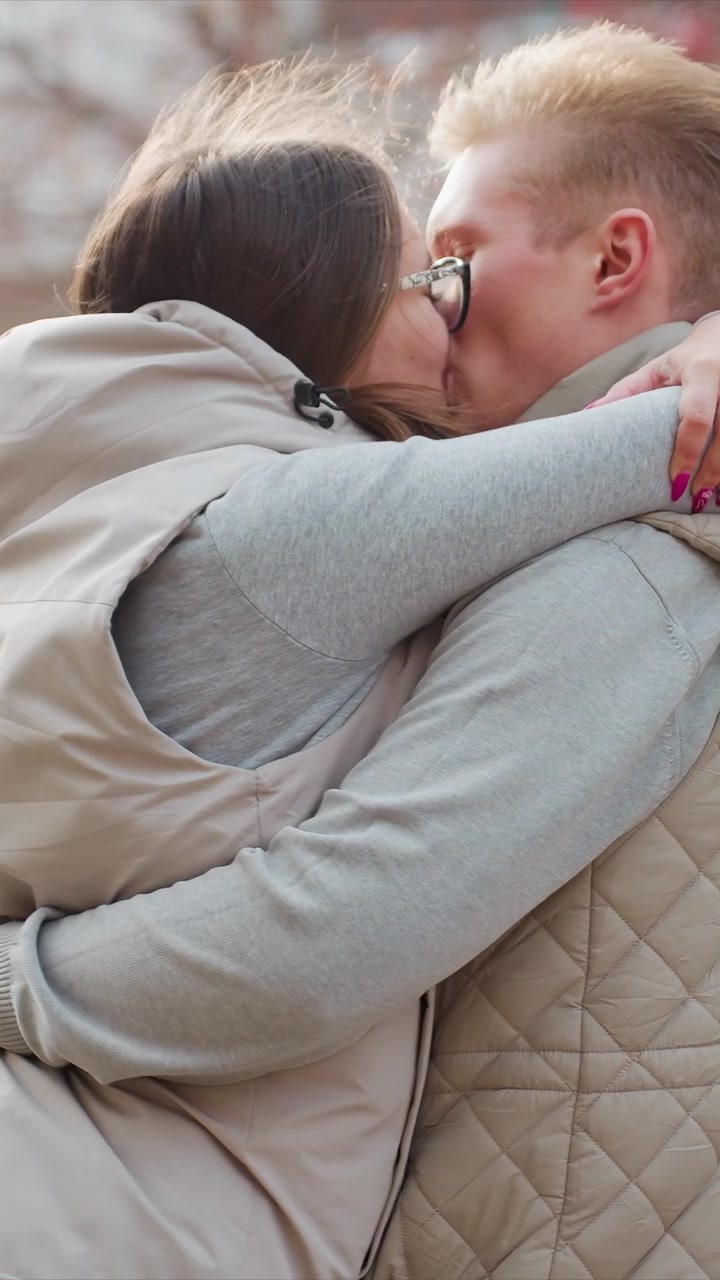 Una donna con gilet beige e felpa con cappuccio grigia ne abbraccia un'altra. Dettagli dell'abbigliamento come tasche del gilet beige, felpa con cappuccio. Momento intimo catturato con enfasi sulla vicinanza fisica.