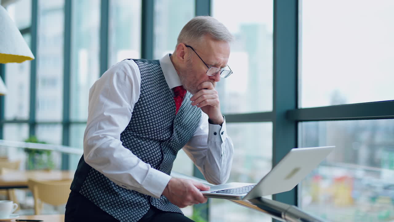 Serious businessman in glasses looking into a laptop. Mature director in elegant suit working on a laptop near the office window.