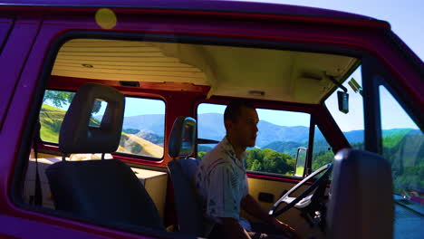 young man with short hair is sittin in an red retro bus in a beautiful mountain landscape and is looking out of the window