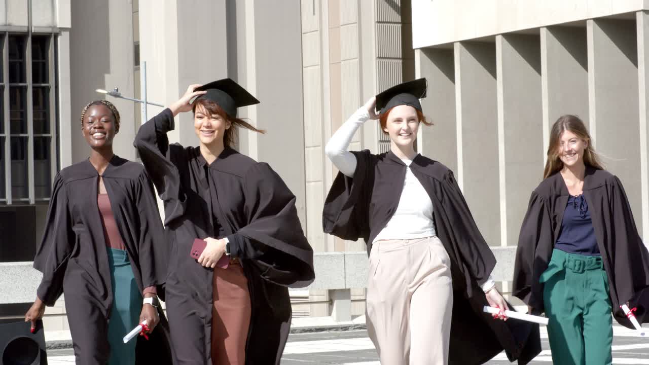 Amiche di diverse etnie camminano attraverso la piazza indossando toche e tocco, tenendo i diplomi, toccandosi i cappelli.