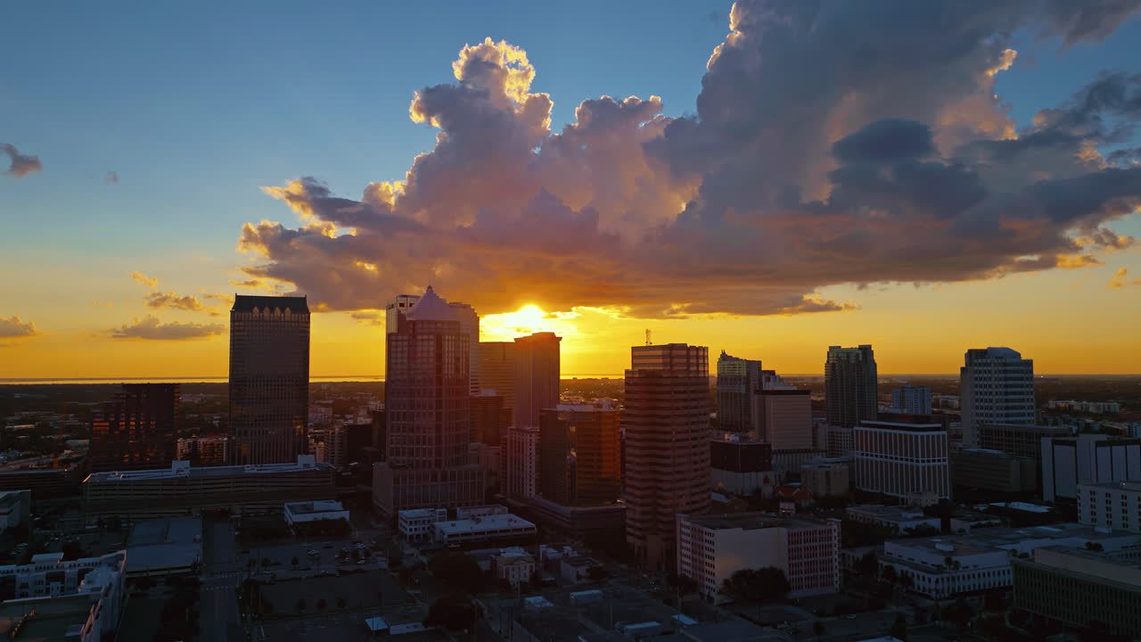 Glowing Tampa skyline at golden hour. A sweeping aerial tracking shot of the city's architecture under a warm, beautiful sunset sky just before the sun drops behind the buildings