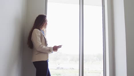 Biracial-businesswoman-standing-using-smartphone-by-window-in-modern-interiors