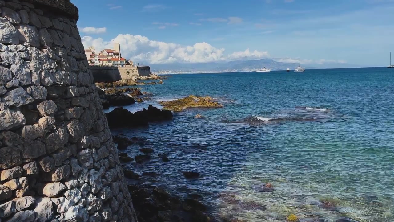 Antichi bastioni in pietra si affacciano su una costa rocciosa mediterranea, acqua turchese, barche lontane e una storica città costiera sotto un cielo azzurro brillante