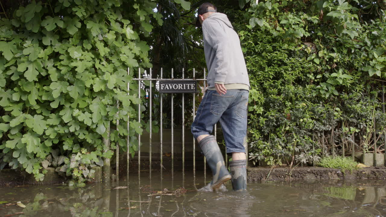 hombre con botas de lluvia entra en la propiedad desde la calle inundada a través de la puerta del jardín