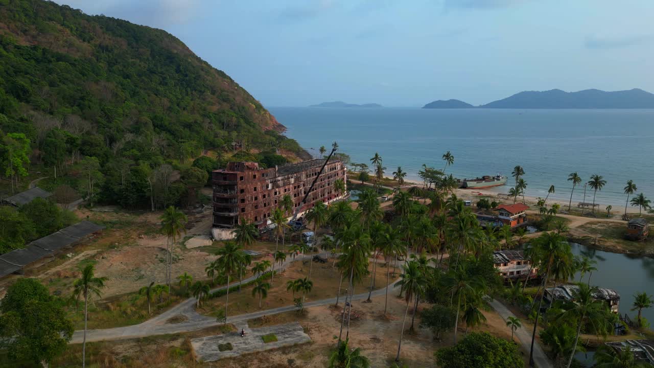abandoned rusty ghost ship on Koh Chang on tropics island bay, Thailand, during sunset, with a crane working on it. Fabulous aerial view flight static tripod hovering drone