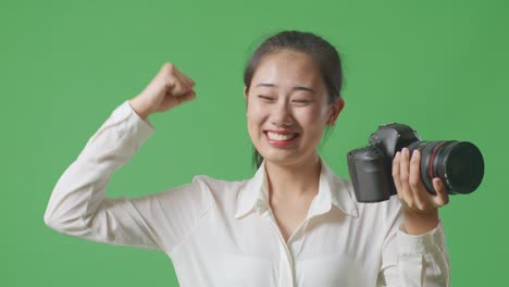 close up of asian photographer using a camera taking pictures and flexing her bicep while standing on green screen background in the studio