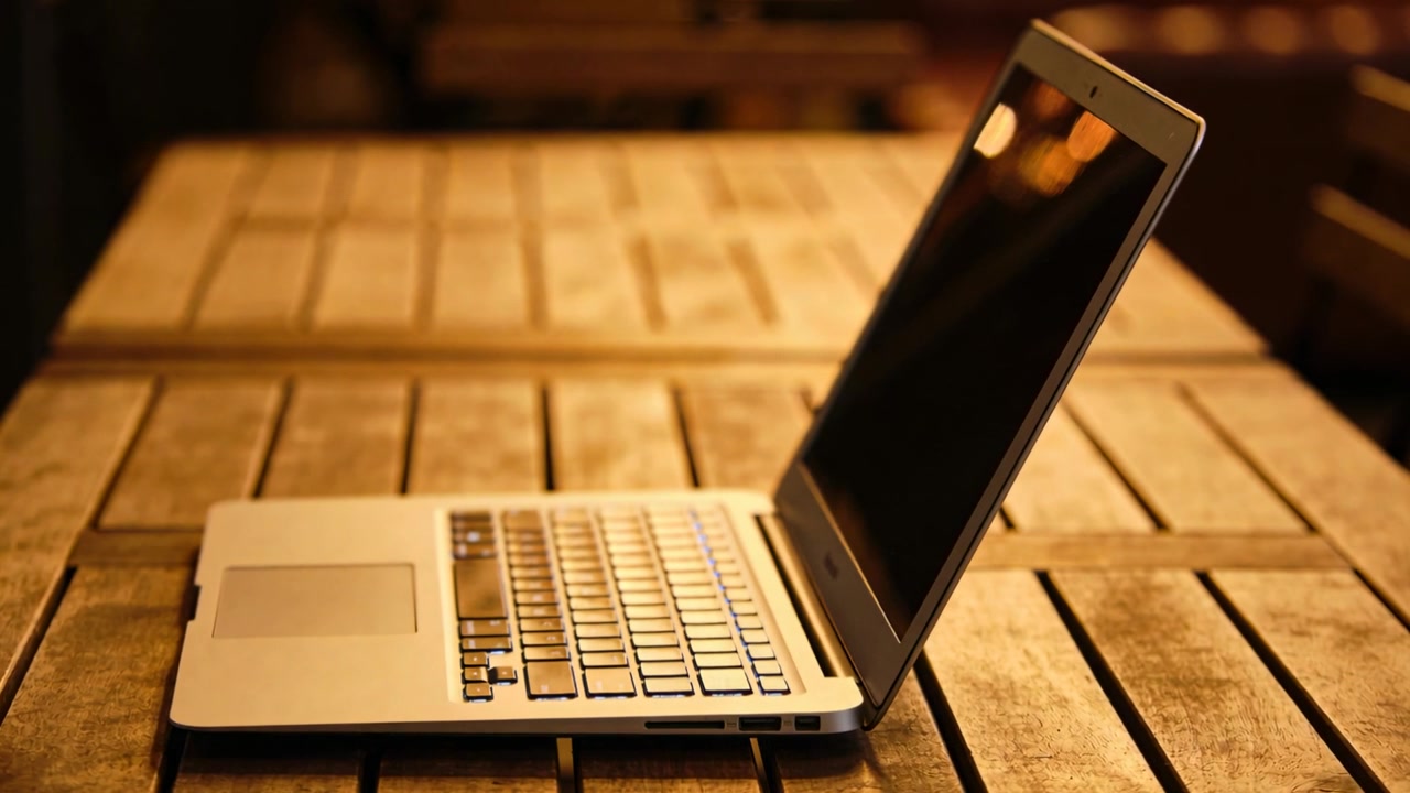 Telecamera che registra un laptop argentato appoggiato su un tavolo a doghe di legno in un caffè accogliente, che cattura riflessi bokeh.