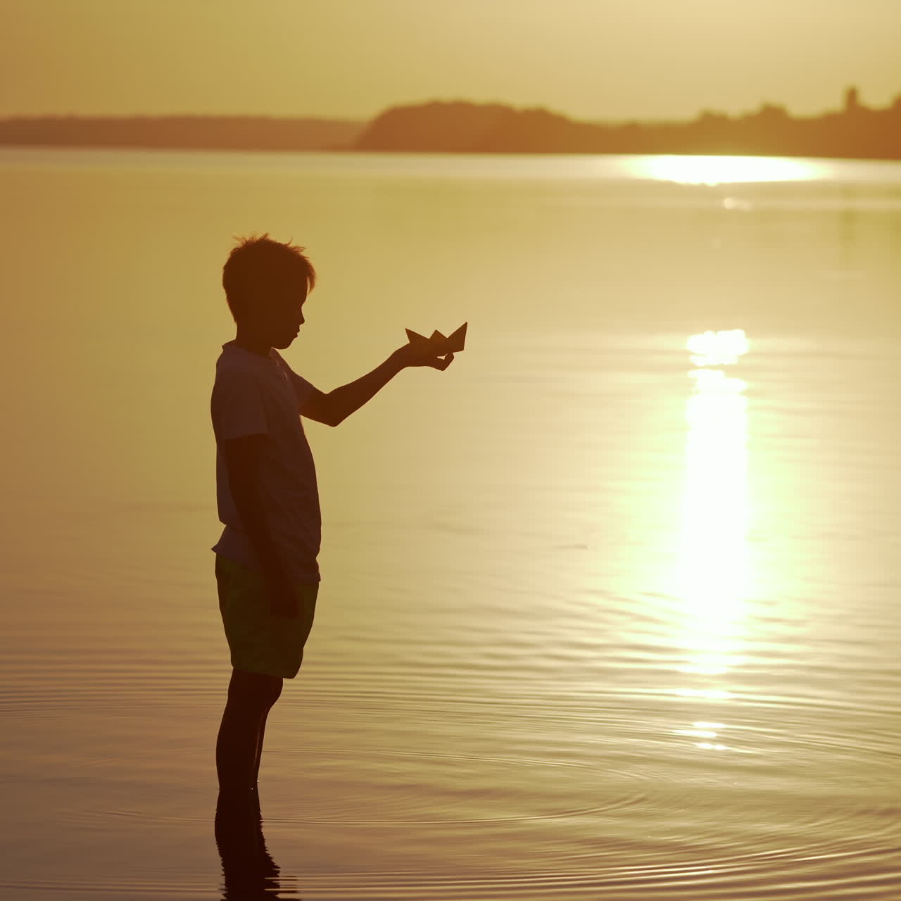 Boy with a Paper Ship is standing in the river. Origami Paper Boat in kid's hands. Beautiful Waves Ripple Background. Travel and nature Concept. Video at sunset.