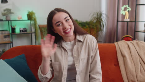 a woman sitting on a couch in her living room, smiling and waving at the camera