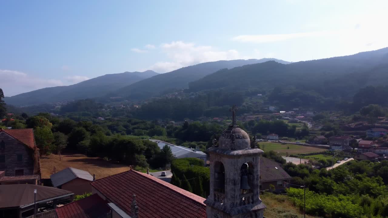 4K drone orbital flight around a stone cross and church tower, showcasing the traditional village houses and lush hills of the Galician countryside, Spain, on a bright, sunny day