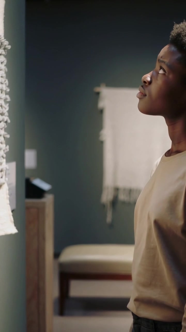 Vertical video: Camera showing museum as teen male moving in viewing textile, tan top, copy space