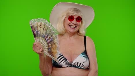 a senior woman in a bikini, hat and sunglasses, smiling and holding a fan