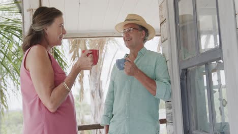 happy senior caucasian couple drinking coffee and talking on porch of beach house, in slow motion