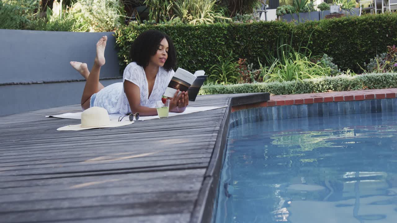 Sunlit African American woman lying on pool deck, reading book, sipping drink, relaxing, copy space