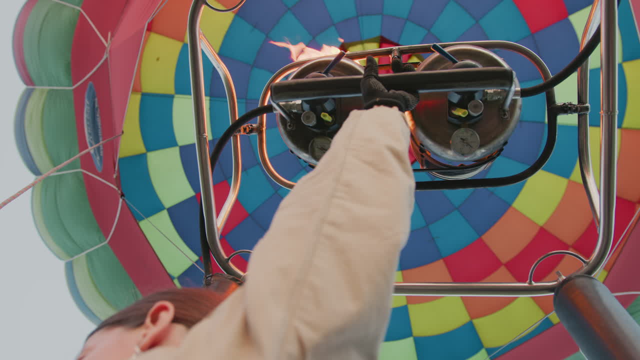 close up of gloved hand adjusting burner controls as flame shoots up into colorful hot air balloon canopy showing patchwork pattern of panels with bright sky in background