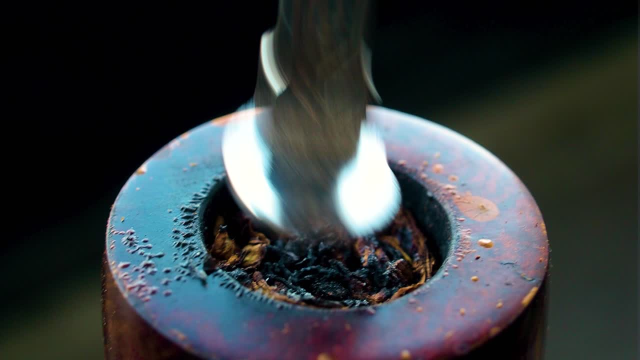 empujando hacia abajo el tabaco con una herramienta de plata dorada para quemarlo mejor y más eficientemente dentro de un agujero de tubería antigua vintage que ha sido dañado por toda la quema colocada en interiores en un ambiente oscuro