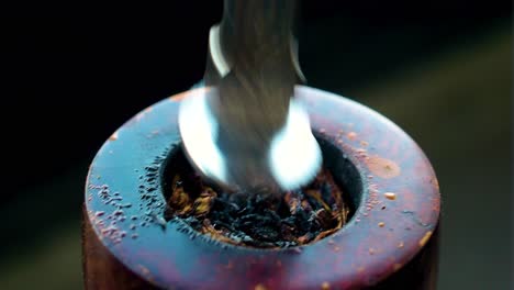 pushing down tobacco with a golden silver tool to burn it better and more efficiently inside a vintage antique pipe hole that has been damaged from all the burning placed indoors in dark environment