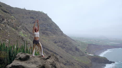 Al-Atardecer,-Una-Joven-Practica-Yoga-En-Una-Costa-Rocosa,-Encarnando-Un-Estilo-De-Vida-Saludable,-Armonía-Y-La-Conexión-Entre-Los-Humanos-Y-La-Naturaleza,-Con-El-Telón-De-Fondo-Del-Océano-Azul