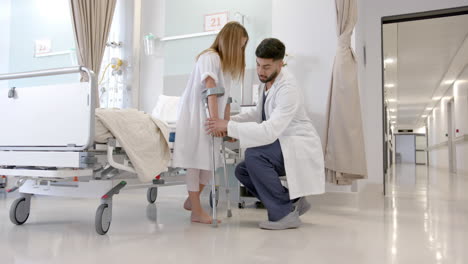 diverse male doctor helping girl patient use crutches in hospital ward, slow motion