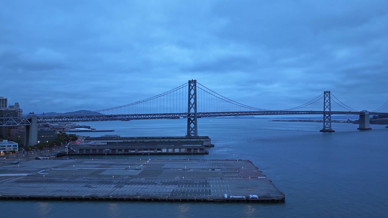 Riprese aeree catturano il San Francisco Bay Bridge al calar della sera, mostrando acqua tranquilla, architettura iconica e una soffusa illuminazione blu sotto un cielo drammatico. Primo piano del lungomare urbano