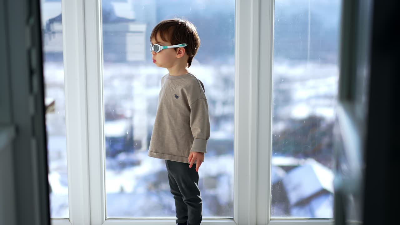 A little boy stands near a window in sunglasses and looks out the window.