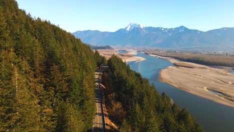 Cinematic-Scene-of-the-Lougheed-Highway-7-running-through-the-forest-in-the-Fraser-Valley-in-the-Lower-Mainland-in-BC,-Canada,-on-a-sunny-day-in-autumn,-cars-driving-snow-capped-mountains,-blue-sky