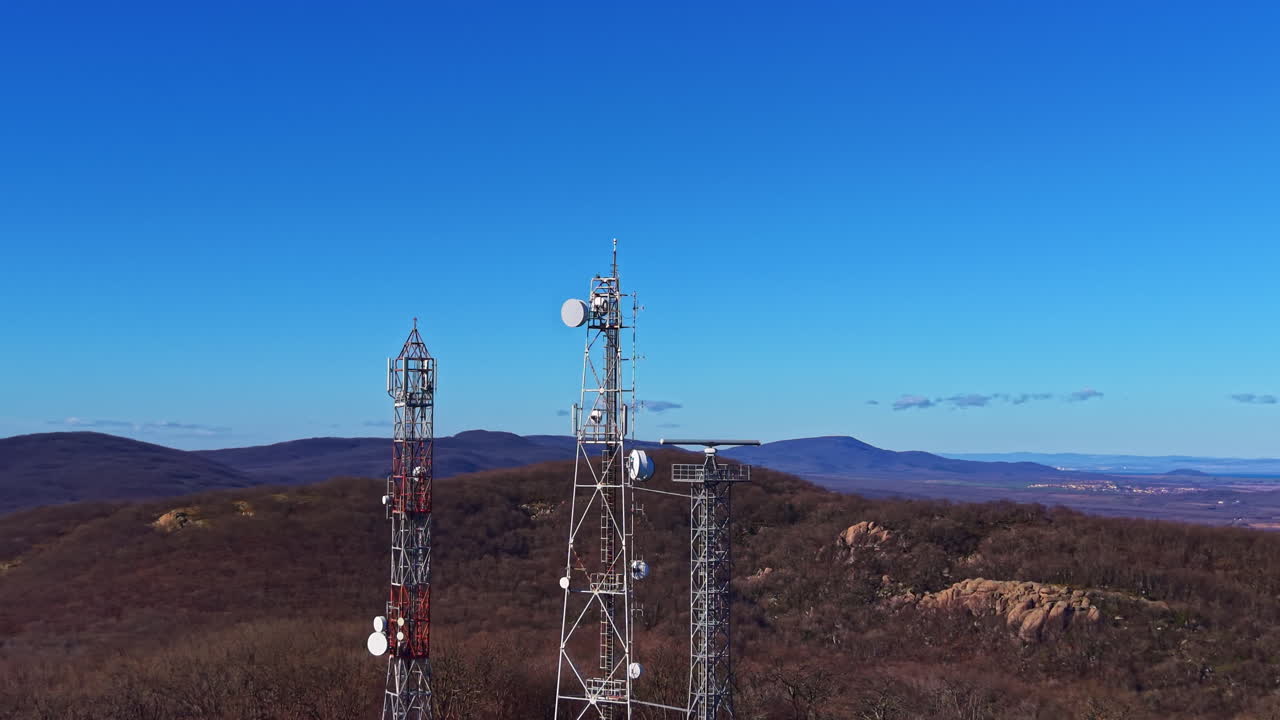 Telecommunication towers stand tall on a hill under a clear blue sky near a mountain range with sparse trees and open land
