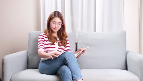 Beautiful-young-Asian-model-flipping-through-picture-magazine-sitting-on-sofa-at-home