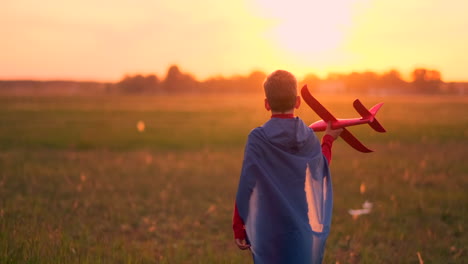 Junge-Läuft-In-Einem-Roten-Regenmantel-Mit-Einem-Flugzeug-In-Der-Hand-Und-Lacht-Bei-Sonnenuntergang-Auf-Der-Sommerwiese.-Er-Stellt-Sich-Vor,-Er-Sei-Ein-Flugzeugpilot,-Der-Mit-Einem-Modellflugzeug-Spielt.