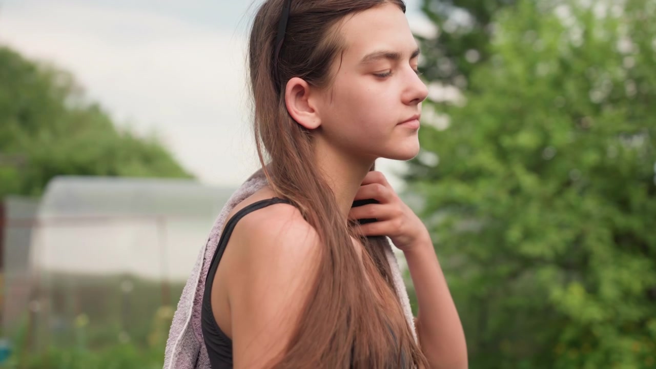 Giovane donna con lunghi capelli castani che indossa un reggiseno sportivo nero e occhiali da sole sulla testa si asciuga dopo l'esercizio. La donna avvolge o sistema un asciugamano viola chiaro intorno alle spalle