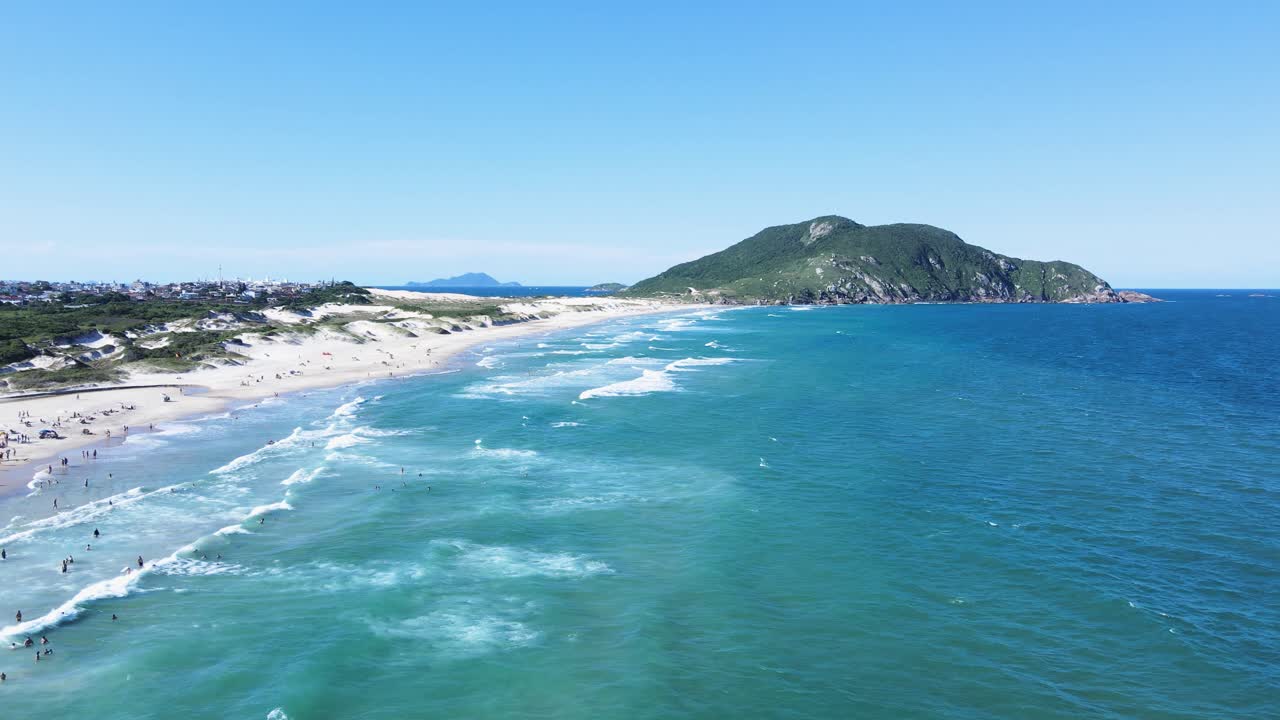 Ampia ripresa aerea cinematografica della spiaggia di Santinho a Florianopolis, Brasile, che mostra le onde turchesi dell'oceano, le dune di sabbia bianca e la montagna Morro dos Ingleses sotto un cielo azzurro e limpido