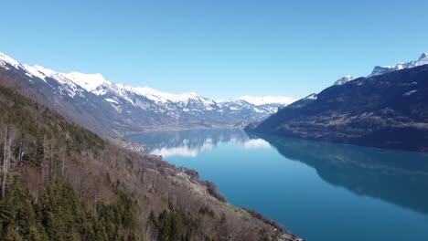 Flight-over-a-mountain-lake-in-Switzerland,-the-snow-covered-alps-are-reflected-in-the-clear-water-like-in-a-mirror,-by-drone