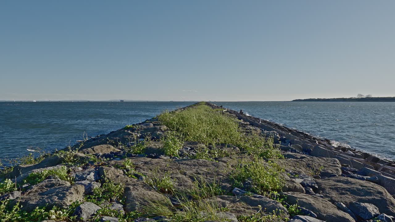 A head-on view of a long, straight, grass-covered rocky jetty or breakwater extending into the bright blue ocean horizon