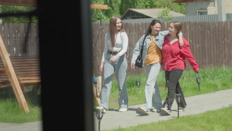three young women walk side by side along garden path, smiling and talking joyfully, one carrying shopping bag, surrounded by wooden fence and bright greenery in sunny outdoor compound setting