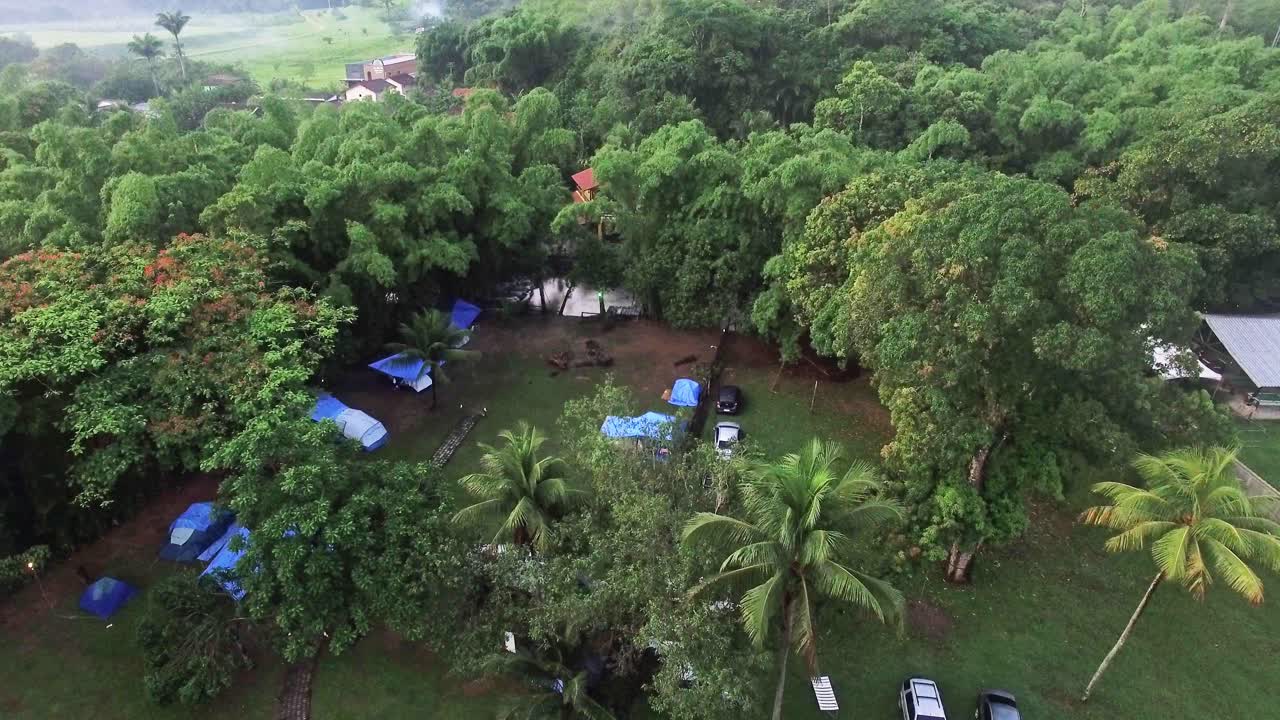 Stunning 4K aerial shot of a campsite with tents, surrounded by lush greenery and trees, beside a small river. The drone moves forward, capturing the beauty of the natural, peaceful camping setting.