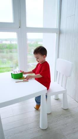 caucasian toddler wearing red t-shirt and blue pants sits at desk playing. focused baby boy plays with educational toy. vertical video.